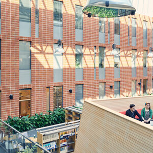 Three people sit in a modern atrium with brick walls, large windows, and hanging lights. Plants and bookshelves are visible.