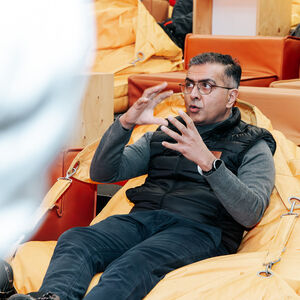 Tarun Sharma, wearing a black vest and grey sweater and sitting on a yellow bean bag chair, is talking and gesturing with their hands.