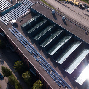 Aerial view of large flat-roof building with glass skylights, solar panels and nearby road with parked vehicles