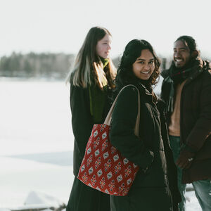 Three friends in winter coats stand smiling outdoors on snow, one with a red patterned tote bag.