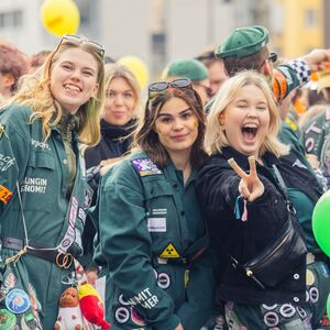 Nuoria aikuisia vihreissä haalarissa ulkoilmajuhlassa, hymyilevät ja pitävät kirkkaita ilmapalloja