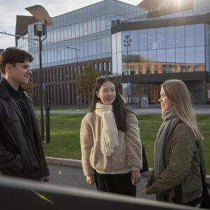 Three students chatting outdoors in autumn clothes in front of a modern glass and brick campus building
