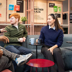 Five young adults chat on sofas in a modern lounge, holding mugs around small red tables.