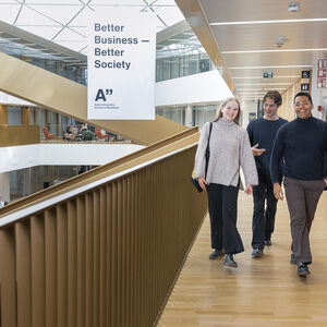 Three students walk in a bright wooden hallway with a large Better Business Better Society banner in the background