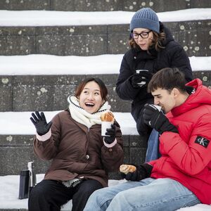 Three friends on snowy steps, dressed in winter coats, laughing and eating pastries and drinking hot drinks