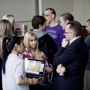 Two business students, one is holding a laptop, discussing with the Professor after his lecture. Another group of students talk in the background.