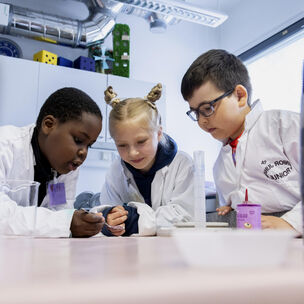 Kolme lasta valkoisissa laboratorio takeissa tekee kokeen luokkahuoneen pöydällä