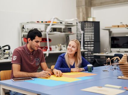 Two students drawing on coloured paper at a table in a workshop space