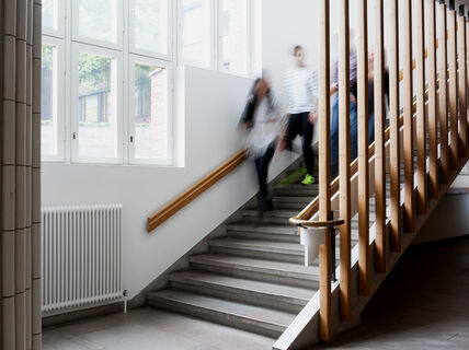 Several people walk down a staircase inside a building with large windows and vertical wooden beams.