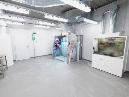 A clean workshop with bright fluorescent lights, spray paint booth, metal sink, white cabinets, and ventilation pipes on ceiling.