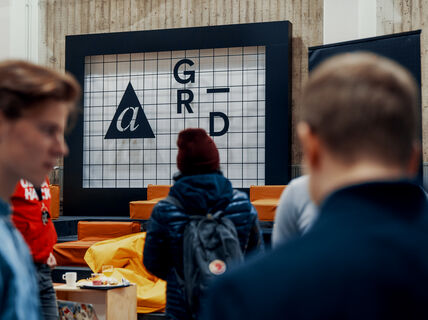 three men talking to each other in the foreground, while another person with back turned looks at the A Grid sign on the background