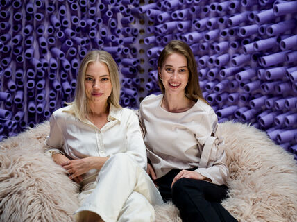 kaksi naista istuu vaalealla karvataljalla violeteista putkista koostuvan taustan edustalla ja katsoo kameraan