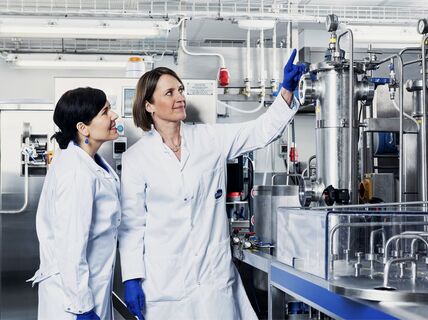 Two lab-coated women in a laboratory looking at pipes.