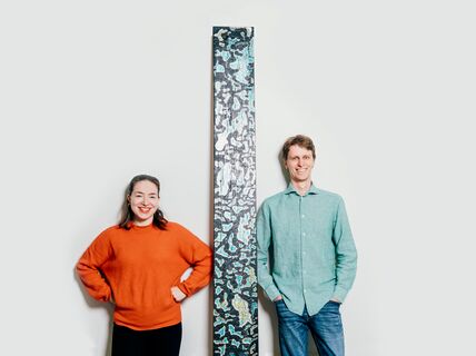 Tohtorikoulutettavat Noora Yau ja Konrad Klockars seisovat vierekkäin valkoista taustaa vasten. Heidän välissään on pystysuora puinen levy, joka on pohjaväriltään musta, mutta pinnassa on kirkkaita sinisiä ja keltaisia kuvioita. Kuva: Jaakko Kahilaniemi.