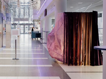 Kuvassa taideinstallaatio SOONI vaaleassa aulatilassa. Teoksen värikkäät tekstiilit liikkuvat teoksesta kantautuvan äänivirran mukana.