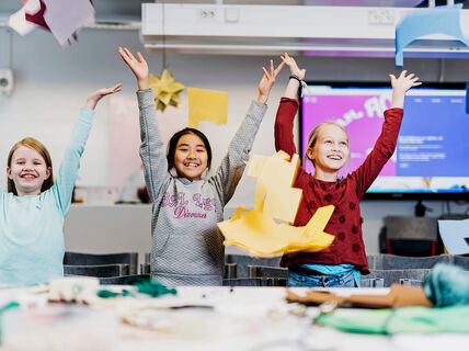 Three children with arms raised in a classroom, surrounded by colourful paper crafts and a monitor in the background.