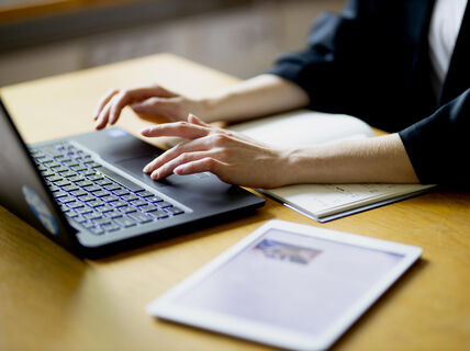 A person typing on a laptop, with notebook and table open, sun shines on the items