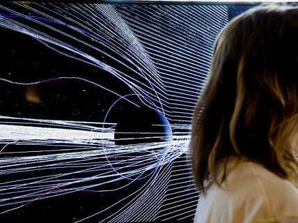 On the right edge a person with shoulder-length hair is moving out of the picture; behind them is a black background with narrow light strings horizontally on it.