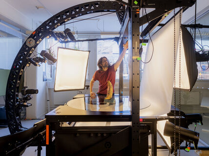 Suureen kaarevaan telineeseen on kiinnitetty kamera, jossa on pitkä objektiivi. Se on kohdistettu keskellä olevaan tasoon, jossa on keltainen keraaminen esine. Pöytätason vieressä on suuri valaisin ja valkokangas sen edessä. Pöydän takana punaiseen t-paitaan pukeutunut henkilö säätää laitteistoa.