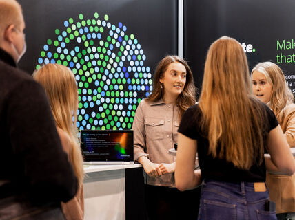 crowd of people are standing in front of black wall with green dots and some text. Deloitte logo on the right-hand upper corner. 