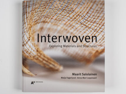 Interwoven-kirjan harmaansävyinen kansi, jossa on kuparilankainen kudos.