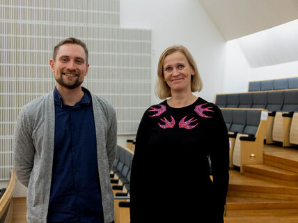 A man and woman in casual wear, dark tones, smiling at the camera inside the Aalto Hall.