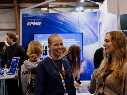 Two women smiling and standing in a crowd. Behing a blue colorful wall with TV screen and KPMG logo on it.