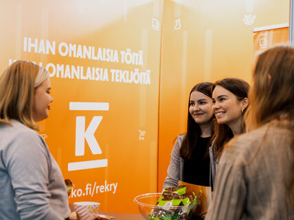 Four people standing infront of orange-colored walls with texts and K group logo. A bowl of candy.