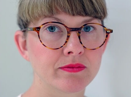 Kuvassa on silmälasipäinen vaaleahiuksinen nainen, jolla on huulipunaa ja vaalea pusero 