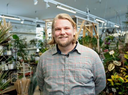 Harmaata ruutupaitaa käyttävä henkilö seisoo kaupassa, joka on täynnä erilaisia kasveja ja puutarhatarvikkeita.