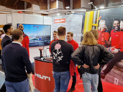 People are standing in front of a wall with huge picture of people with red shirts sitting on a coffee break and a Hilti logo. People are talking with eachoterh.