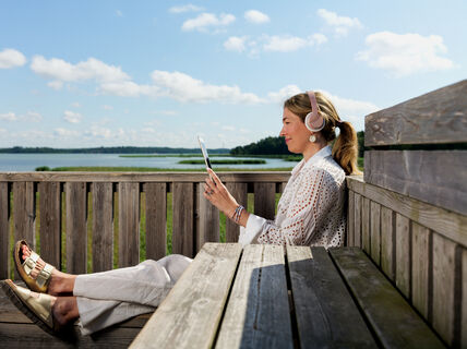 A person with headphones sits on a wooden bench using a tablet, with a lake and cloudy sky in the background.