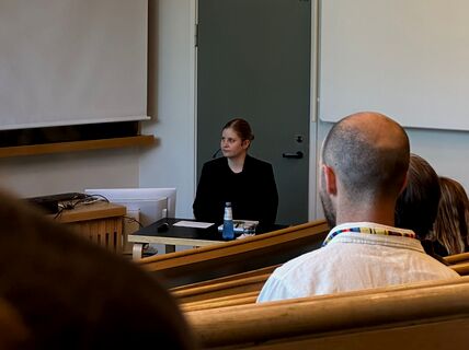 Nainen tummassa puvussa väitöstilaisuudessa luentosalin edessä