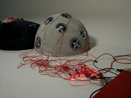 Henkilö makaa lattialla pää betonista tehdyn kuvun sisällä, kuvussa on lukuisia johtoja