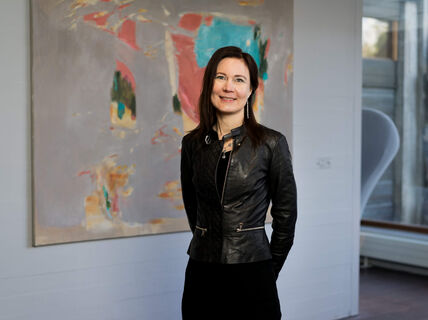 Nainen seisoo taulun edessä ja katsoo hymyillen kameraan.