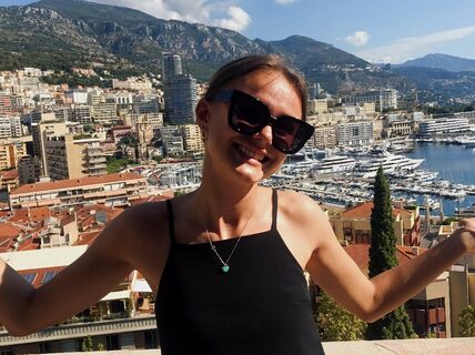 A woman is standing outside with her hands spread out. A town and sea is seen in the backgrond.