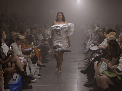A model walking the catwalk shows off an outfit created by a fashion designer, while audience sits watching the fashion show