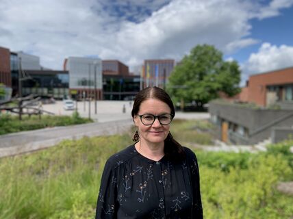 Dark-haired woman in dark dress standing outdoors with Otaniemi campus on a summer day