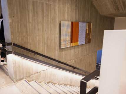 Betoninen portaikko rakennuksen sisällä, seinällä värikäs maalaus, portaissa näkymästä poistumassa henkilö.