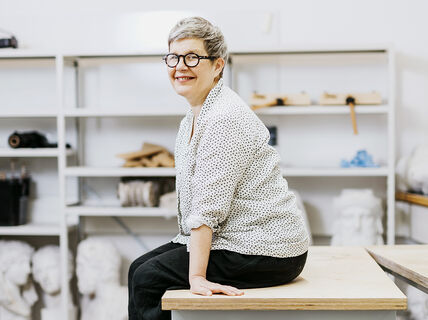 Marja Rastas istumassa pöydällä työpajassa, taustalla näkyy hyllykkö sekä kipsipatsaita.