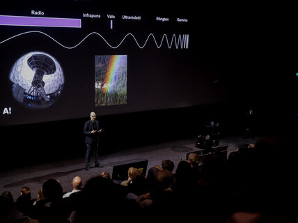A person is presenting to an audience with a large screen showing a wave diagram and a rainbow.