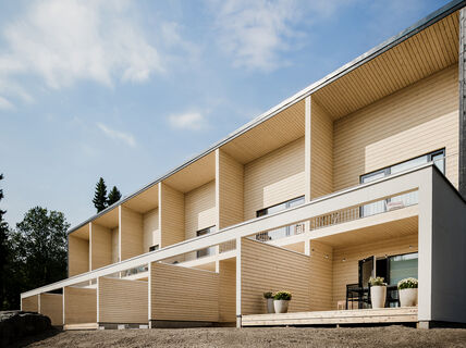 Modern row house with wooden cladding, large terraces, and potted plants outside, under a partly cloudy sky.