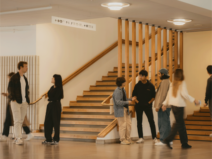 A group of people talking and walking near a wooden staircase indoors, with handrails and overhead lights visible.