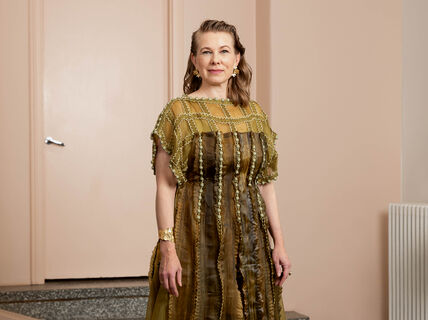 A person stands indoors wearing a green dress with vertical lines and textured details. They have accessories on their wrist and ears.