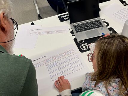 Two people working on experiment sheets at a table with a laptop and water bottle.