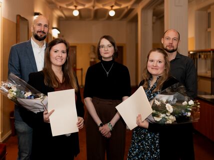 Group of people indoors with two holding bouquets and certificates, standing in a hallway.