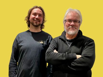 Two people standing against a yellow background; one in a dark shirt, the other in a dark jacket with arms crossed.