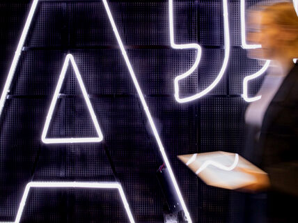 A person holding a tablet stands in front of a large illuminated letter 'A' on a dark background.