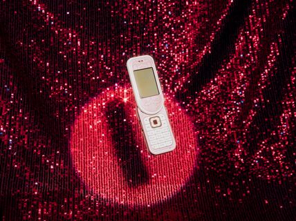 A white flip phone with a screen open lies on a sparkling red sequined fabric.