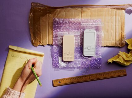 Two mobile phones in plastic bags on bubble wrap and a torn cardboard box, notepad, pencil, and ruler on a purple surface.